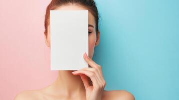 Woman hides face behind a white card on pastel background. Beauty and skincare concept with copy space on a pink and blue split. photo