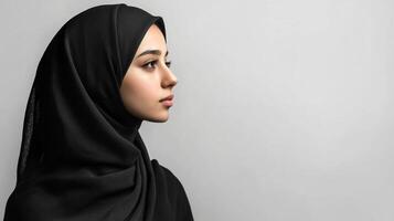 Portrait of woman in dark hijab, side profile against a plain white background. Minimalistic composition emphasizes form and expression. photo