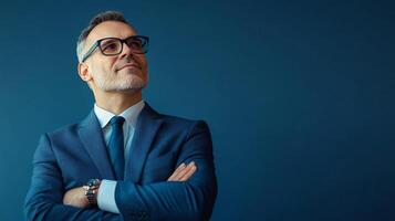Confident businessman in a blue suit stands with arms crossed, looking upward with a hopeful expression against a solid backdrop. photo