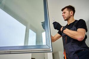 Skilled worker wearing gloves uses screwdriver to fix window frame, home renovation tasks. Professional repairman adjusting white pvc windows in modern building photo