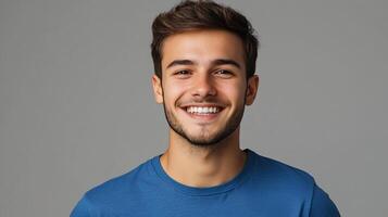 A smiling young man in a blue shirt is pictured against a neutral gray background. He appears happy and approachable in the portrait. photo