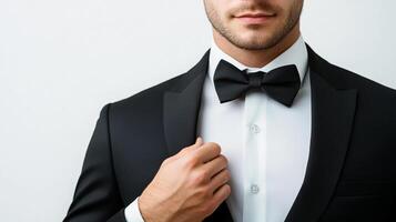 A suited man poses elegantly with a bowtie. He is dressed in a suit, shirt and bowtie in front of a plain background and the photo cropped.