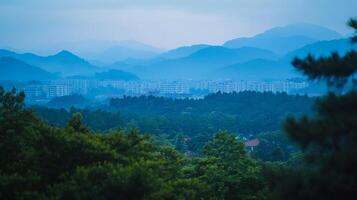 Landscape featuring lush greenery and a skyline of buildings set against a backdrop of hazy, blue-toned mountains on a serene, overcast day. photo