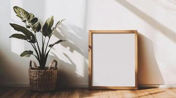 Indoor scene with a leafy plant in a woven basket beside a blank picture frame against a white wall with shadows on a wood floor. photo