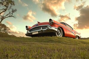 A vintage American car parked in a vast field, with the golden hues of a sunset casting a nostalgic and serene atmosphere photo