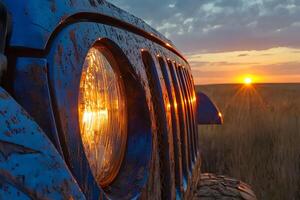 A close-up shot of a blue off-road vehicle parked during sunset, with the golden hues of the setting sun illuminating the vehicle and casting long shadows on the ground photo