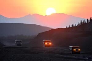 Heavy trucks navigate a mining site at sunset, showcasing the silhouette of mountains against a colorful sky photo