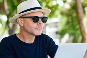 Man working outdoors on a laptop while wearing sunglasses and a hat in a green environment during daylight hours photo