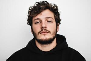 Young man with curly hair and beard wearing a black hoodie poses against a plain white background in a neutral expression photo