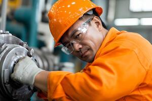 A worker in an orange safety suit and hard hat focuses on maintaining a large machine in a busy industrial setting. photo