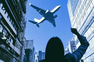 A silhouette of a woman waves towards a flying airplane against a bright blue sky. Surrounding skyscrapers create an urban backdrop, emphasizing the contrast of movement and stillness. photo