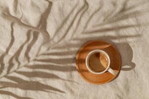 Summer day scene made with cup of coffee and tropical palm tree leaf shadow on sand color linen background. Sun and shadows. Minimal summer coffee concept. Flat lay style. Top view. photo