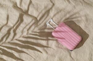 Summer day scene with tropical palm tree leaf shadow and pastel pink baggage on sand color linen background. Minimal stylish travel concept. Trendy summertime adventure idea. Flat lay, top view. photo