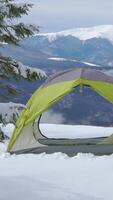A bright green tent is set up in a snowy landscape, surrounded by mountains. The clear sky enhances the serene atmosphere of this winter camping location. Vertical footage video