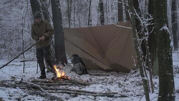 A man is preparing a fire while standing in a snowy forest. A campfire crackles in front of a makeshift shelter in a serene snowy forest. video