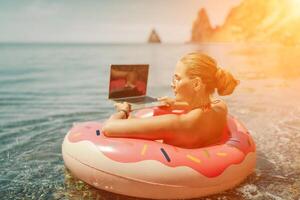 Woman laptop sea. Freelancer woman in sunglases floating on an inflatable big pink donut with a laptop in the sea. People summer vacation rest lifestyle concept photo