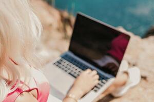 A woman is sitting on a rock with a laptop open in front of her. She is typing on the keyboard and she is focused on her work. Concept of productivity and concentration. photo