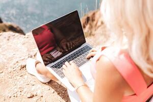 A woman is sitting on a rock with a laptop open in front of her. She is typing on the keyboard and she is focused on her work. Concept of productivity and concentration. photo