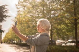 A woman is taking a picture of herself with her cell phone. She is wearing a gray jacket and scarf. The scene is set in a city with trees and cars in the background. photo