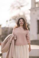 A woman wearing a pink sweater and white skirt is smiling and holding a brown bag. The scene has a warm and cheerful mood, with the woman looking happy and confident. photo
