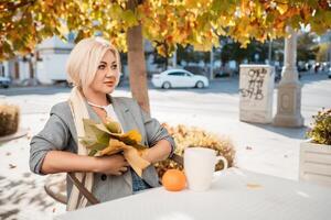 A blonde woman sits at a table with a cup of coffee and a stack of papers. She is wearing a gray jacket and a necklace. The scene is set in a park with trees and benches. photo