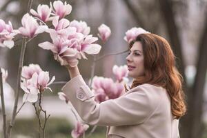 Woman magnolia flowers, surrounded by blossoming trees., hair down, wearing a light coat. Captured during spring, showcasing natural beauty and seasonal change. photo