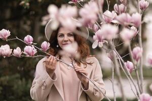 Woman magnolia flowers, surrounded by blossoming trees., hair down, white hat, wearing a light coat. Captured during spring, showcasing natural beauty and seasonal change. photo