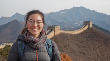 Woman smiling on the Great Wall, wearing glasses, a scarf, and a gray coat, with the Wall and mountains in the background. photo
