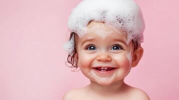 Adorable toddler covered in bubbly foam, big smile, and sparkling blue eyes, enjoying bath time against a soft pink background. photo