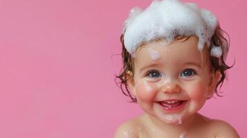 A joyful baby smiles with suds on their head, set against a solid pink backdrop. Glistening skin and bright eyes add to the charm. photo
