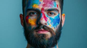 Man with colorful paint on his face in a studio. His beard is dark and he stares intently at the camera with an introspective look. photo