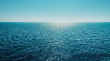 A calm ocean under a clear sky, the horizon blending seamlessly. Sunlight reflects on the water, creating a shimmering surface. photo