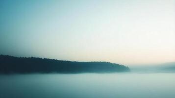 A tranquil landscape featuring a dense forest silhouette emerging from a layer of ethereal mist under a soft, gradient sky. photo