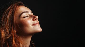 Radiant redhead basks in serenity, eyes closed, a cascade of auburn hair, freckles and black backdrop creates visual harmony. photo