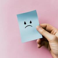 hand holding blue sticky note with sad face emoji against pink background conveys feelings of sadness and disappointment photo