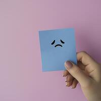 hand holding blue sticky note with sad face emoji against pink background conveys feelings of sadness and reflection photo