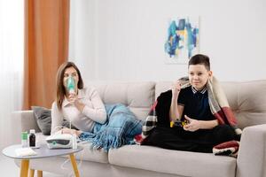 Woman holding a mask nebulizer and inhaling fumes medication into lungs. Sick lady inhaling through inhaler mask at home and funny boy with gamepad playing game at home near her photo