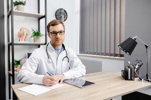 Young doctor wearing glasses and white uniform with stethoscope, speaking and consulting patient online, looking at camera, making call, sitting at table in office. photo