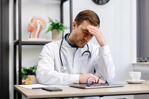 Doctor man wearing medical uniform sitting on his workplace tired holding his head feeling fatigue and headache. Stress and frustration concept. photo