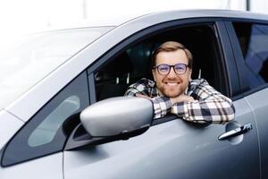 Man customer sitting in car salon and smiling, choosing auto and wanting to buy new automobile in showroom dealership store. Car sales concept photo