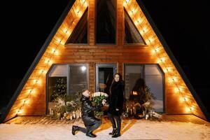 Man kneeling and offering white roses to woman in front of a wooden house at night photo