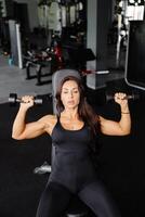 Young fit woman practicing dumbbell press while sitting on a bench. Slim female in sportswear exercises with weights in a gym. photo