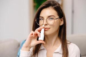 Young woman wearing casual clothing, using a nasal spray to facilitate breathing, sitting on a sofa in a home interior. photo