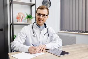 Doctor wearing glasses and white uniform with stethoscope, speaking and consulting patient online, looking at camera, making call, sitting at table in office. photo