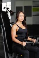 Young woman practicing dumbbell press while sitting on a bench. Slim female in sportswear exercises with weights in a gym photo