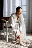 Bride wearing white suit and veil tying elegant high heels sitting on chair in hotel room photo