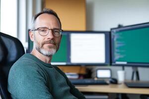 A man with glasses sitting in front of two computer monitors photo