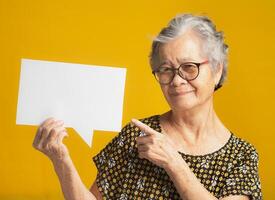 Senior woman holds and points to a blank speech bubble and looks at the camera with a smile photo