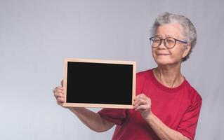 Senior woman holding a mini blackboard and looking at the camera with a smile while standing on a gray background. Space for text. photo