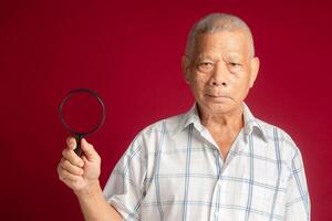 Senior man holding a magnifying glass and looking at the camera while standing on a red background photo